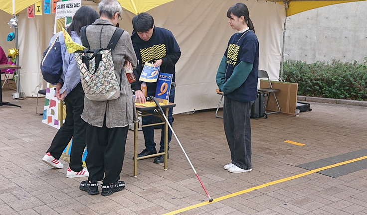 ココテープのブース。製品サンプルが乗った机を挟んで、スタッフが女性と男性のペアに説明している。