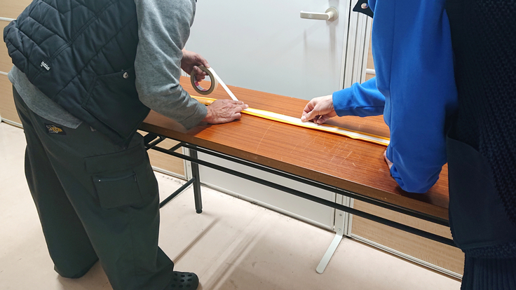 長机の上にココテープ裏面の上にして置き、二人がかりで両面テープを貼り付けている写真