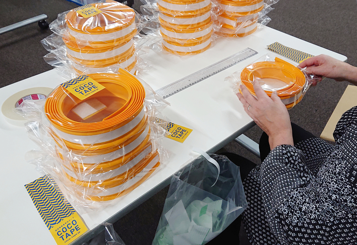 テーブルの上でココテープの袋にシール貼りをしている写真。作業場所の周りにはシール貼り付け済みのココテープが積まれている。