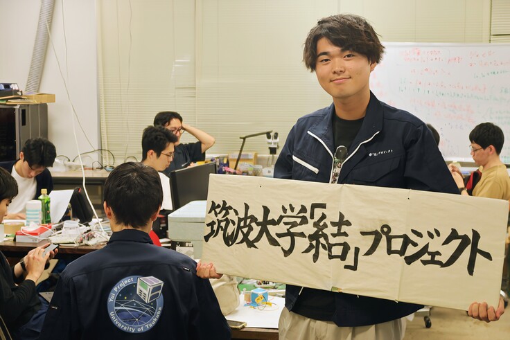 通信系と電源系のミーティングを背景に、ゴール達成を喜ぶ学生代表。