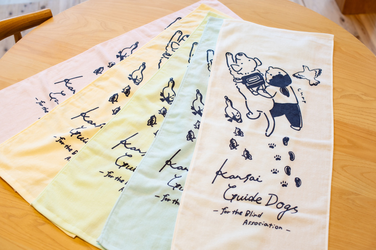 白、ピンク、水色、黄色、黄緑色のタオルに盲導犬と歩くユーザー、カモ、鳩がデザインされている。